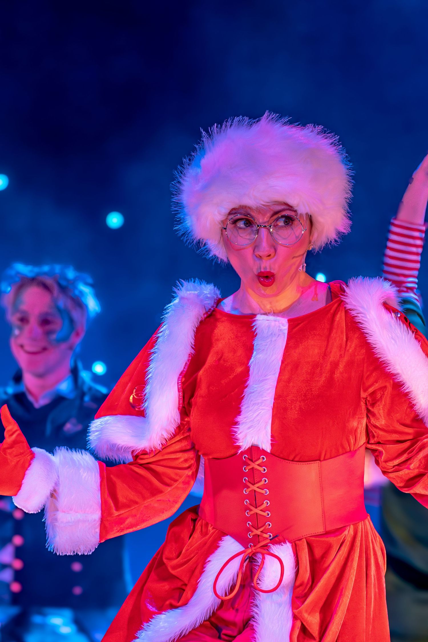 Holiday show performer in red costume and white fur hat expressing surprise under colorful lights.