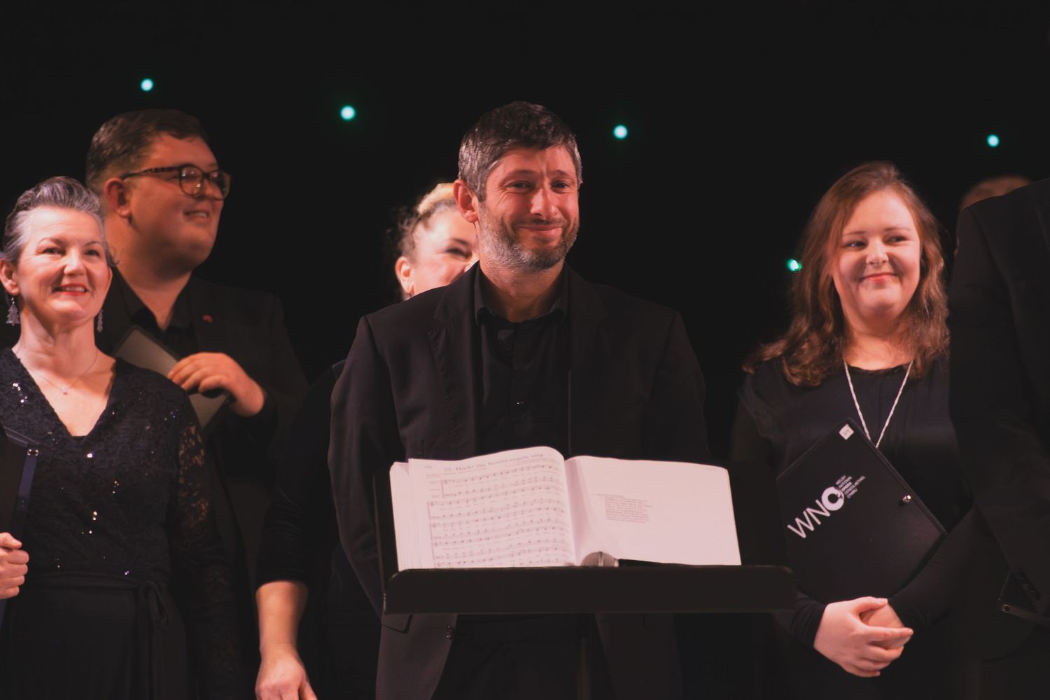 Performers holding sheet music during a choral concert with dramatic lighting.