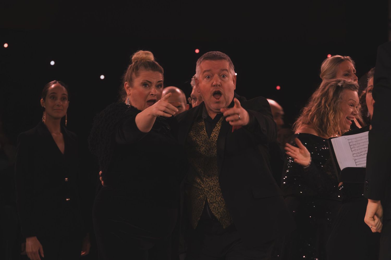 Choir members in black attire sing from sheet music during an evening concert.