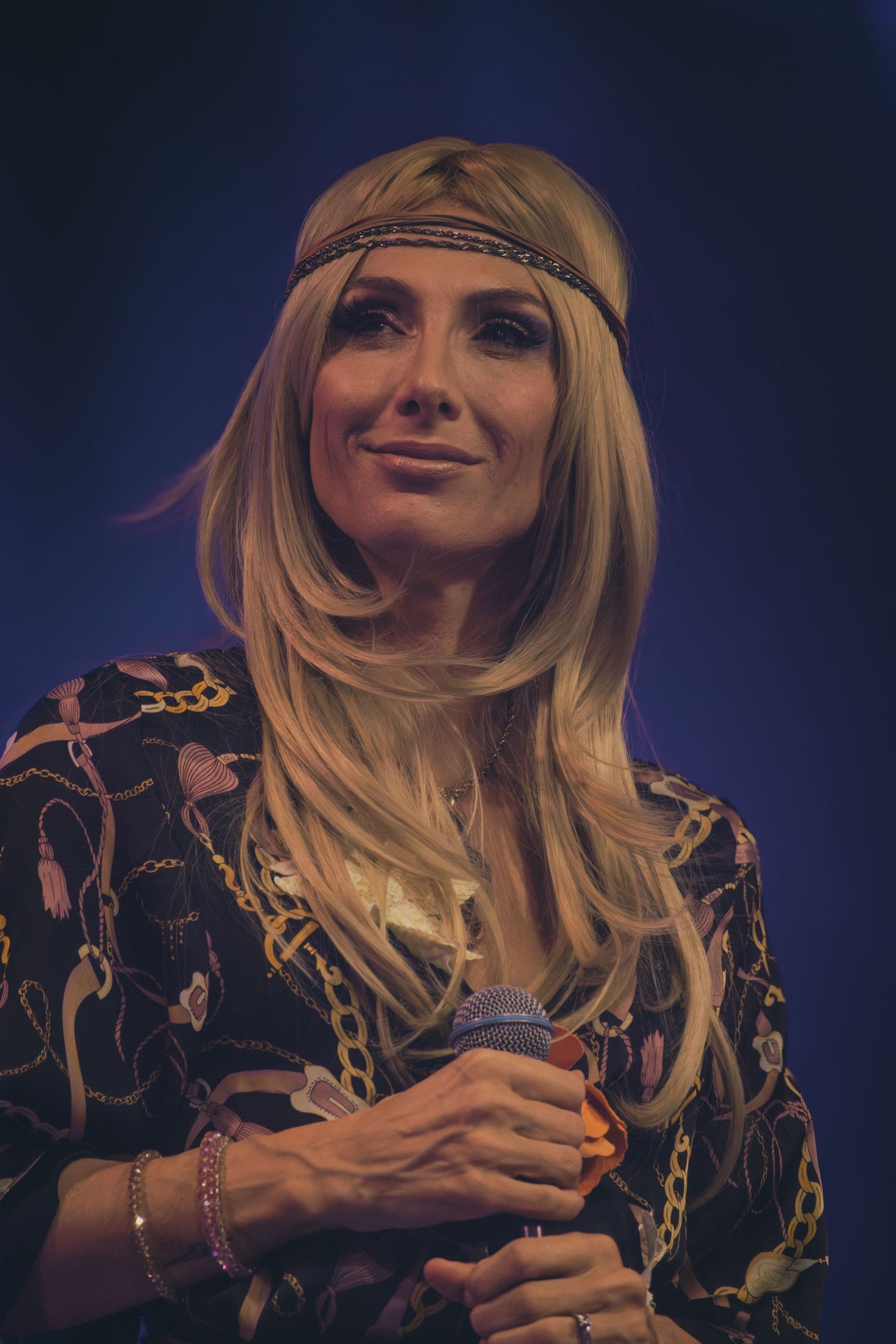 A performer in a boho-style outfit with a headband sings on stage under blue lighting.