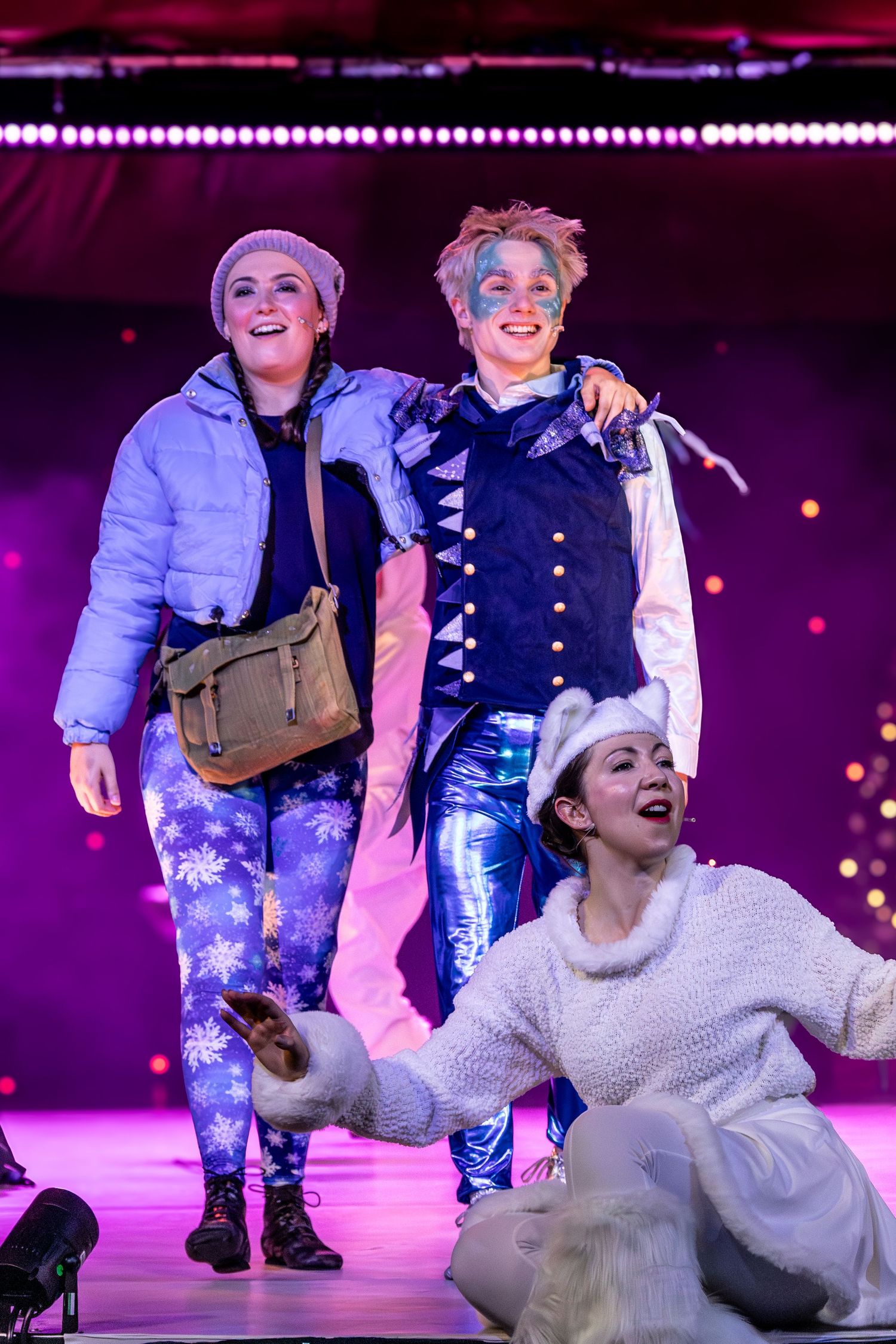 Performers in colorful costumes dance energetically on a purple-lit stage during a theatrical production.