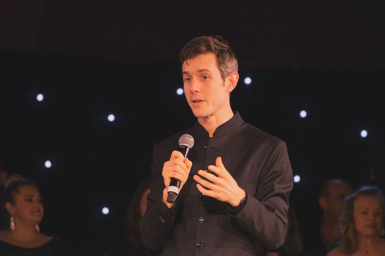 A performer speaks into a microphone on a dimly lit stage with blue lights visible in the background.