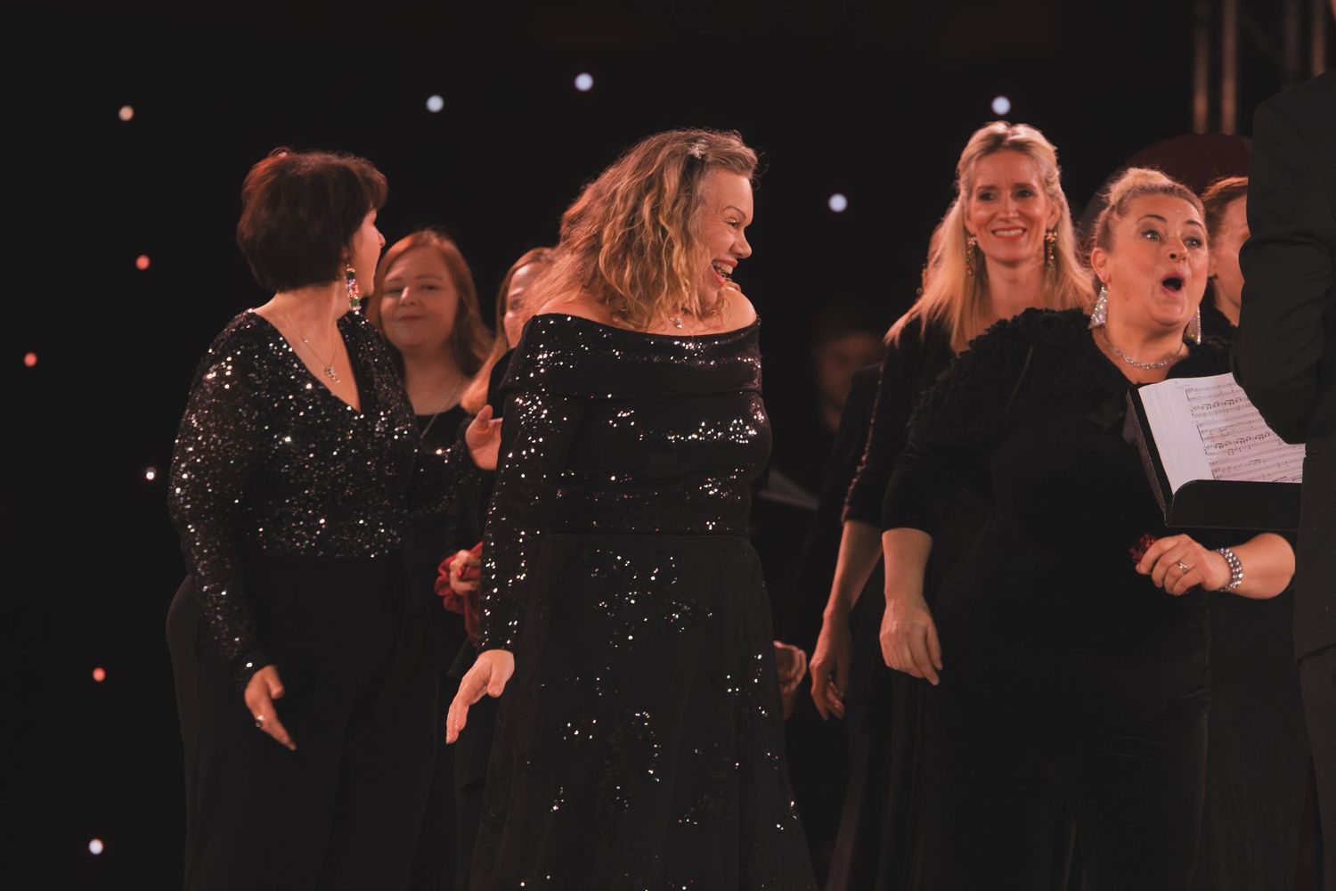 Choir members in sparkly outfits performing on stage with dramatic backlighting.