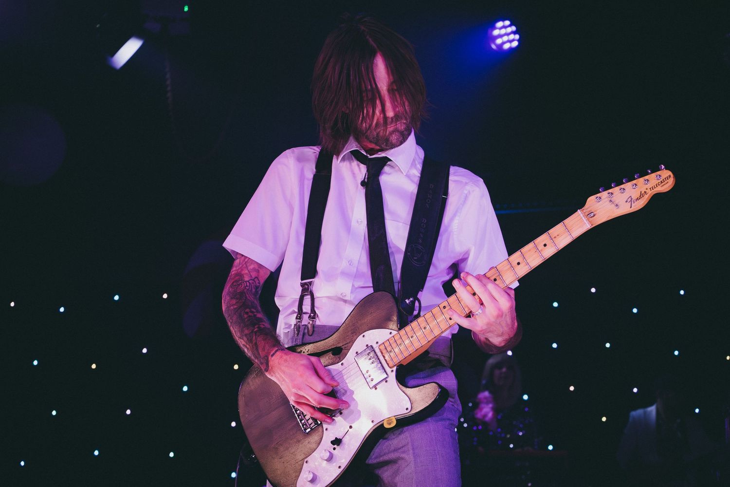 Guitarist performs on stage wearing formal attire under blue stage lights.