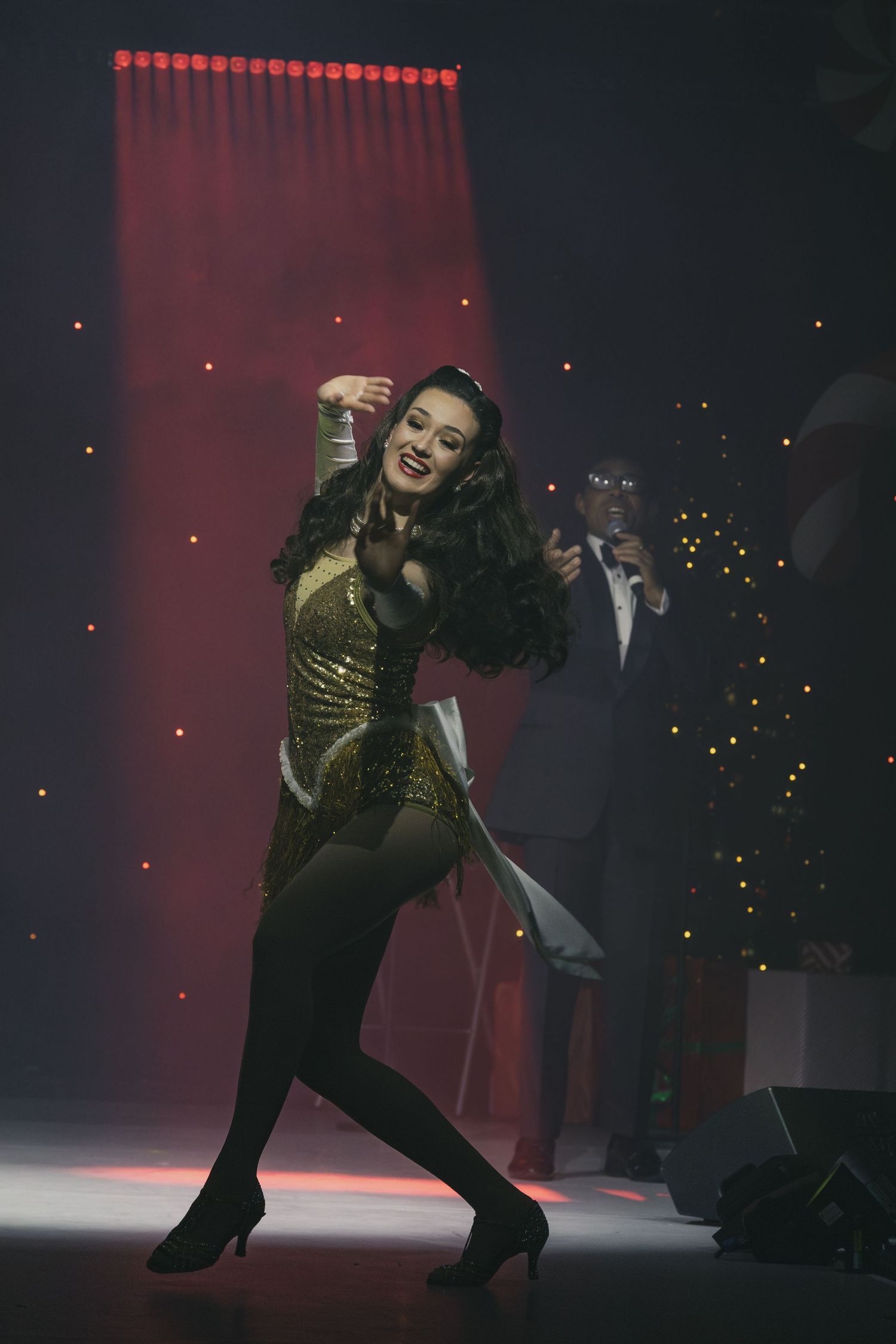 A performer in a gold outfit dances energetically on a dimly lit stage with red lighting effects.