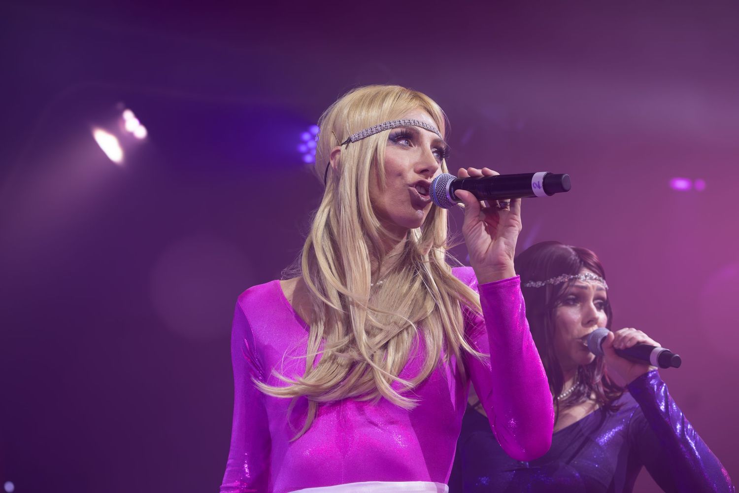 Performer in pink outfit singing passionately during a concert with vibrant purple stage lighting.