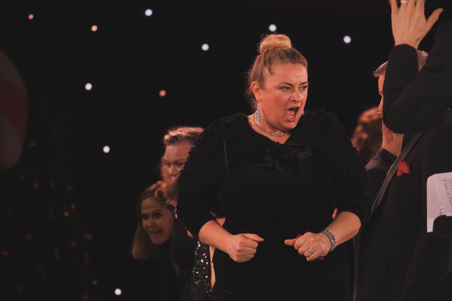 Performers in black attire singing on stage with spotlights overhead.