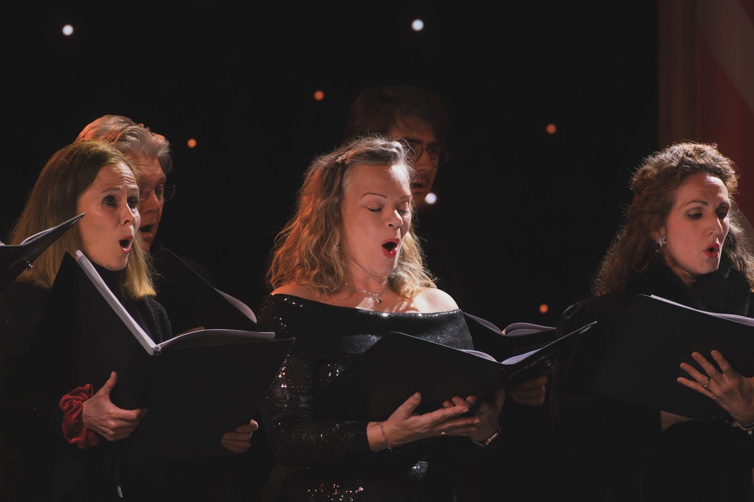 Performers in black clothing hold sheet music while singing in a darkened concert hall.