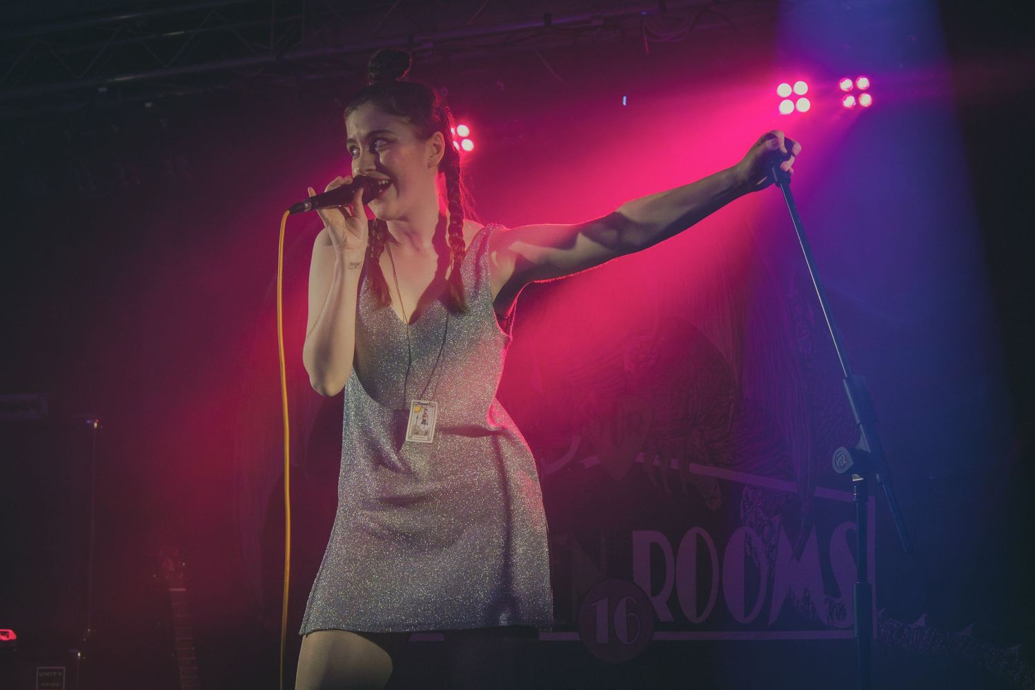 Concert performer in gray tank top singing on stage with dramatic pink and purple lighting effects.