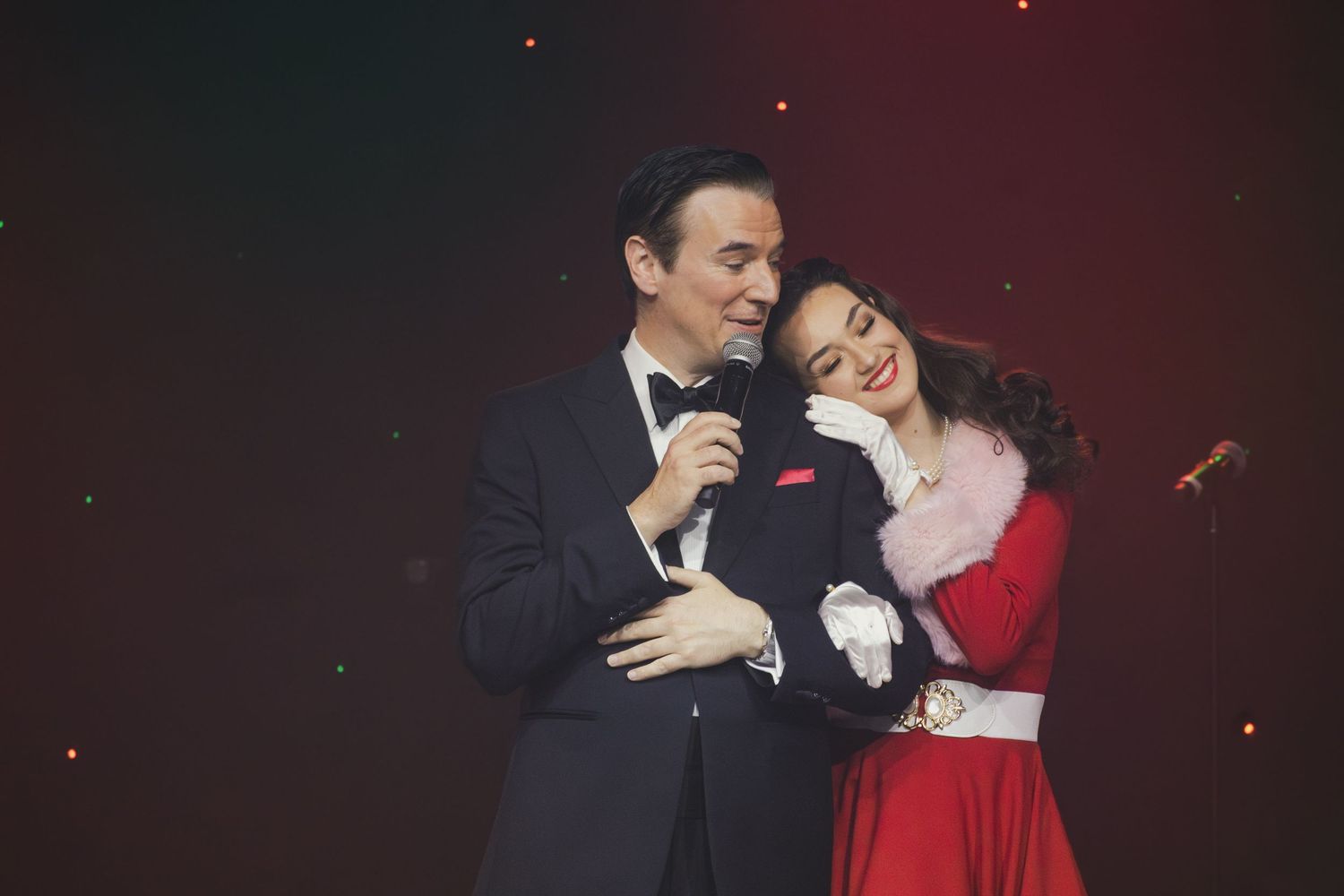 Performers in formal attire sing together on stage with dramatic red lighting and sparkles in the background.