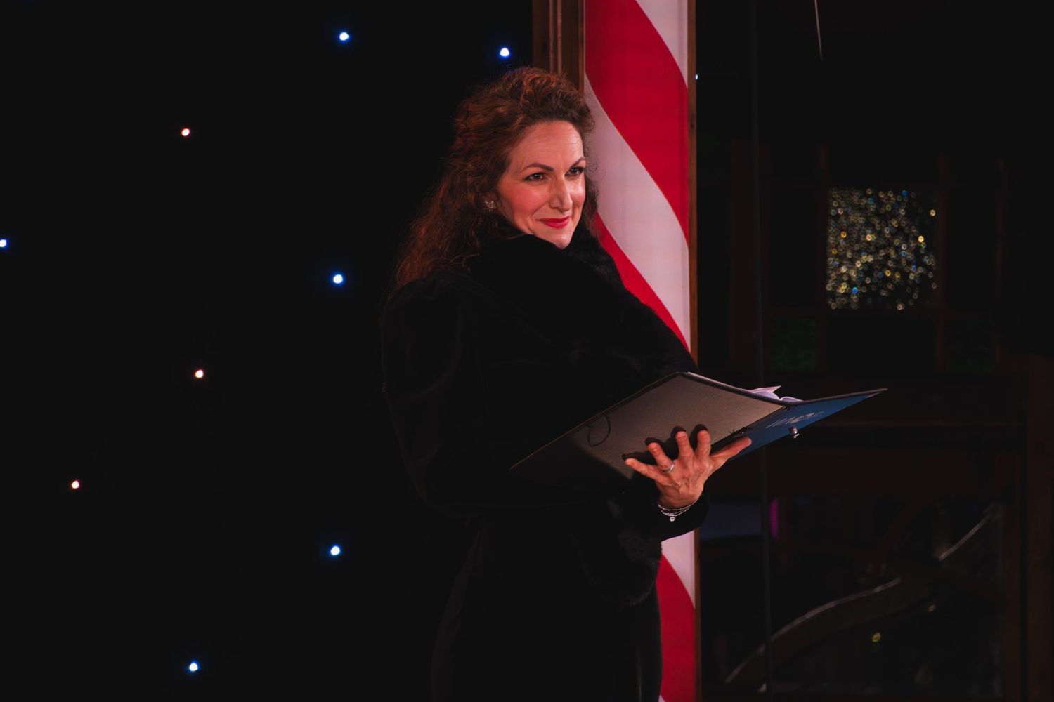 Performer in black dress sings emotively while holding a music folder against dark backdrop.