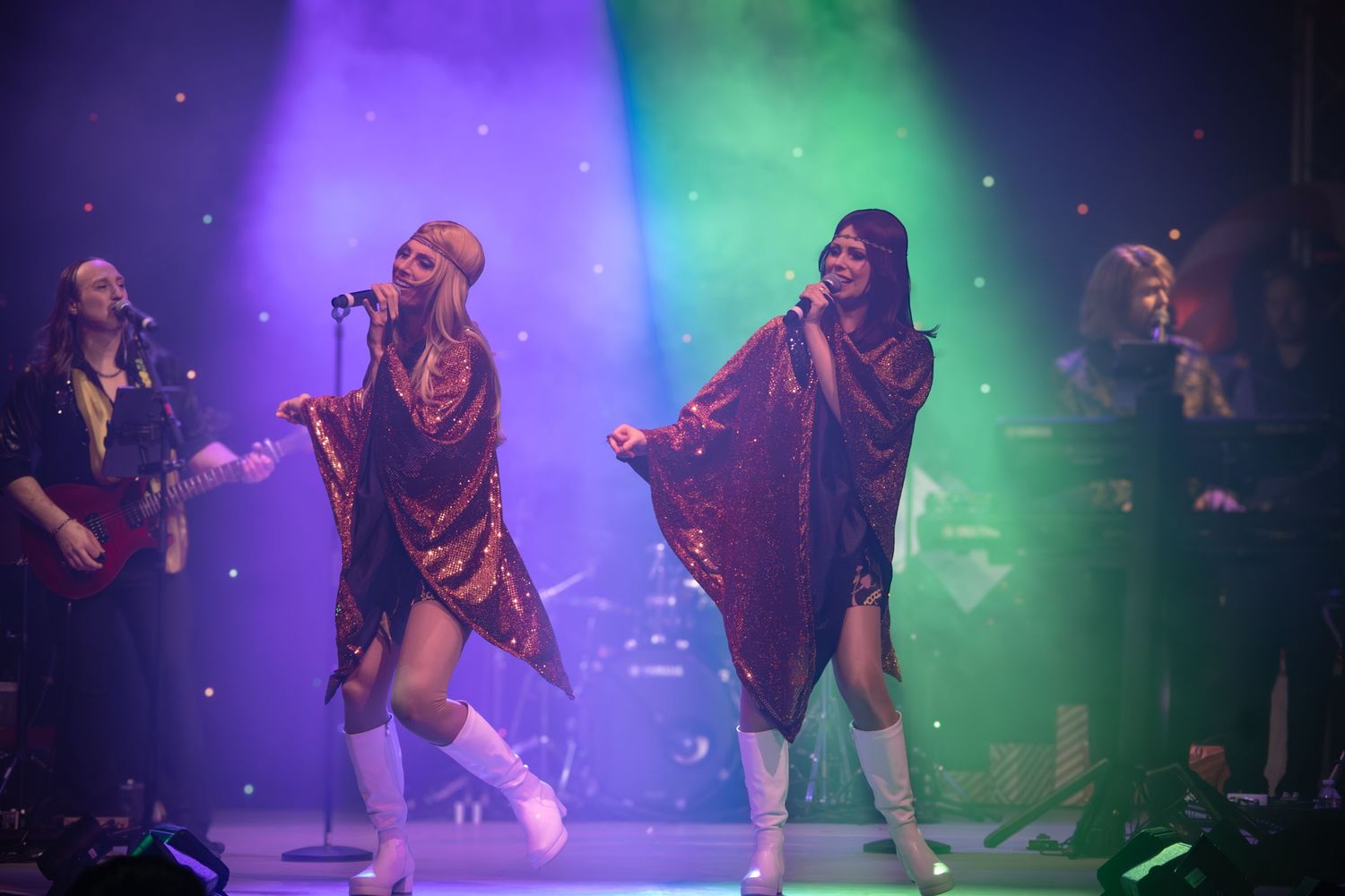 Performers in sparkly outfits dance energetically on stage under purple and green lighting effects.