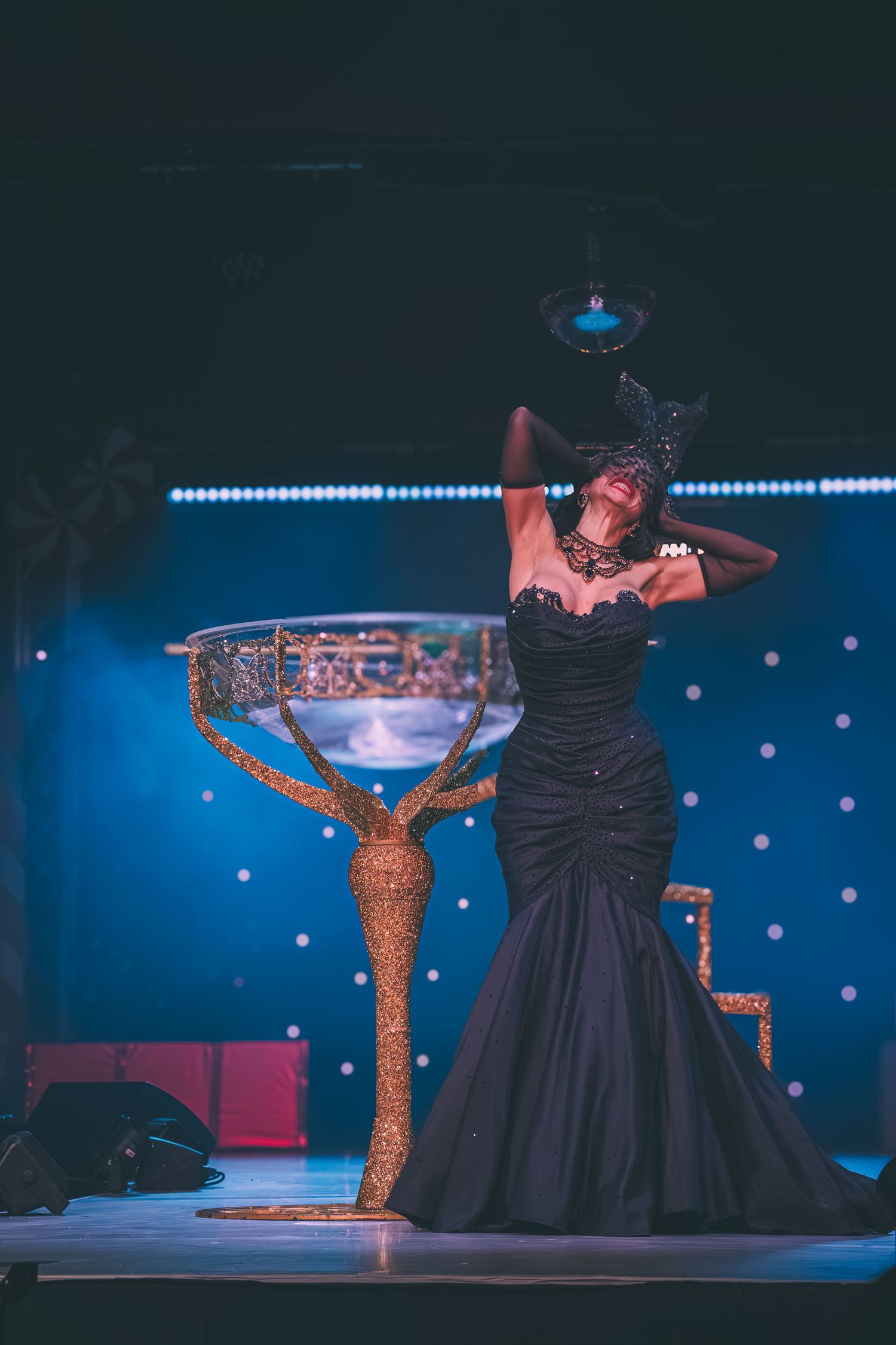 Dancers in elegant black dresses perform on stage with glittering gold props and blue lighting effects.