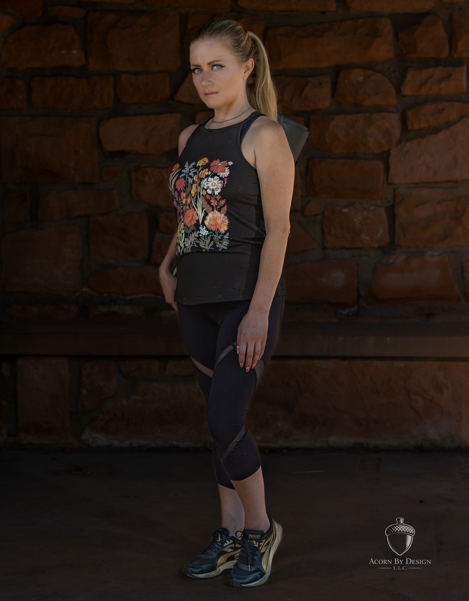 Woman in black floral tank top and cropped leggings poses against stone wall in athletic wear.