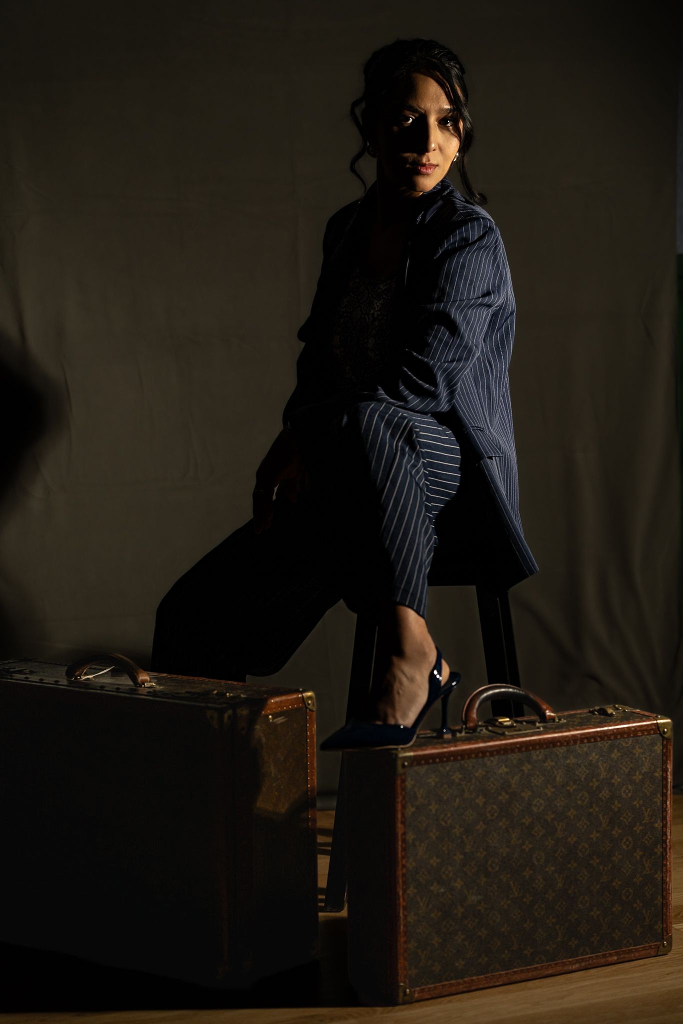 Woman in blue pinstripe suit posing with vintage Louis Vuitton luggage in dramatic studio lighting.