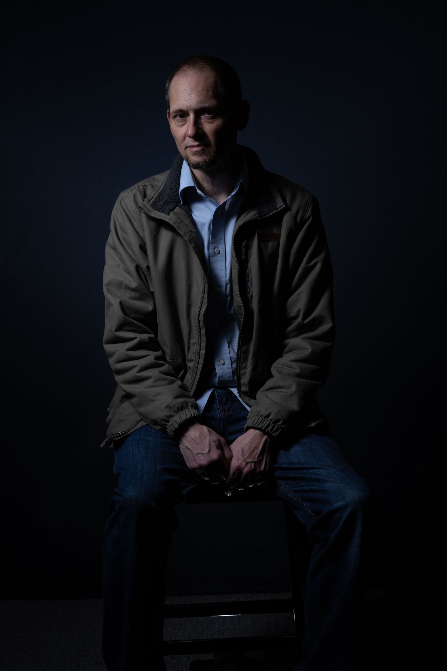 Low-key portrait of person sitting on chair in dark setting wearing casual attire.