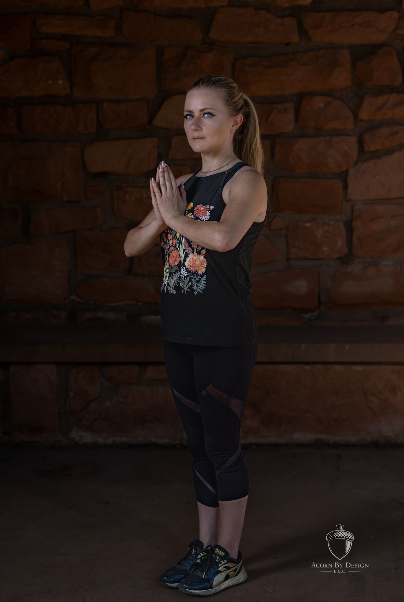 Woman in black floral tank and leggings holds prayer pose before rustic stone wall in fitness photo.