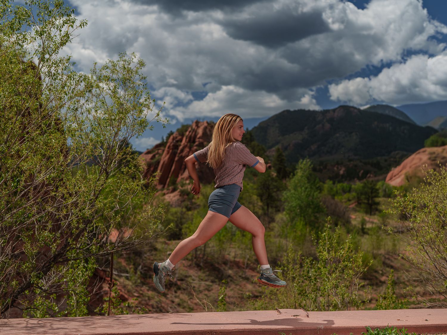 A young blonde girl leaps energetically on a trail with red rock formations and mountains under cloudy skies in Colorado.