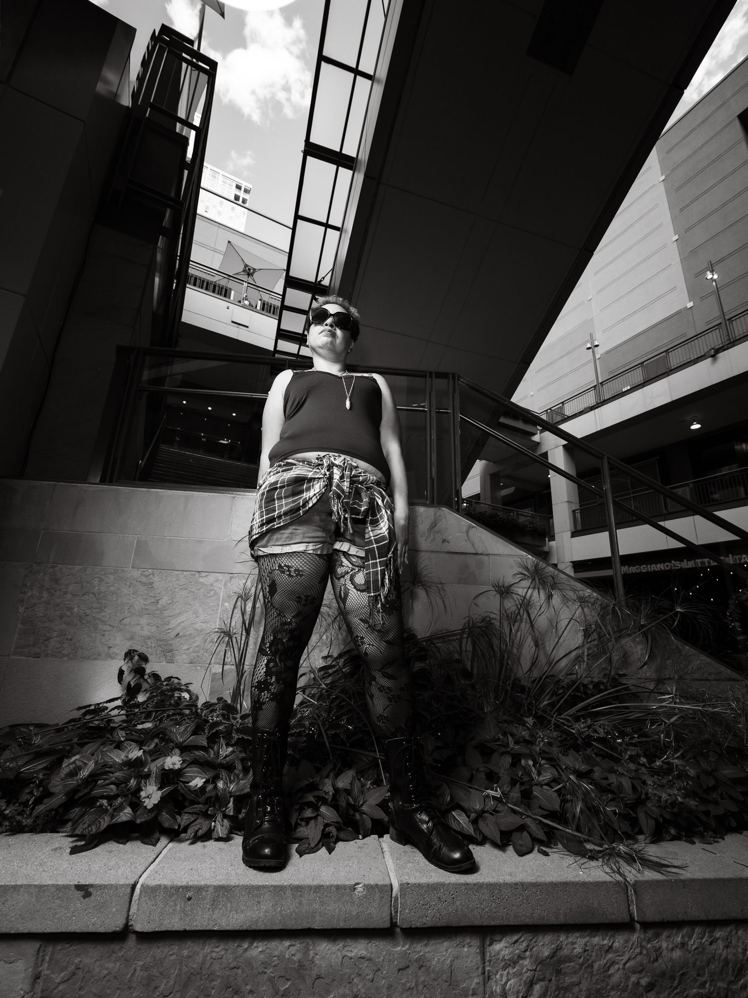Black and white photo of a person in plaid shorts and sunglasses standing among plants outside a modern building.