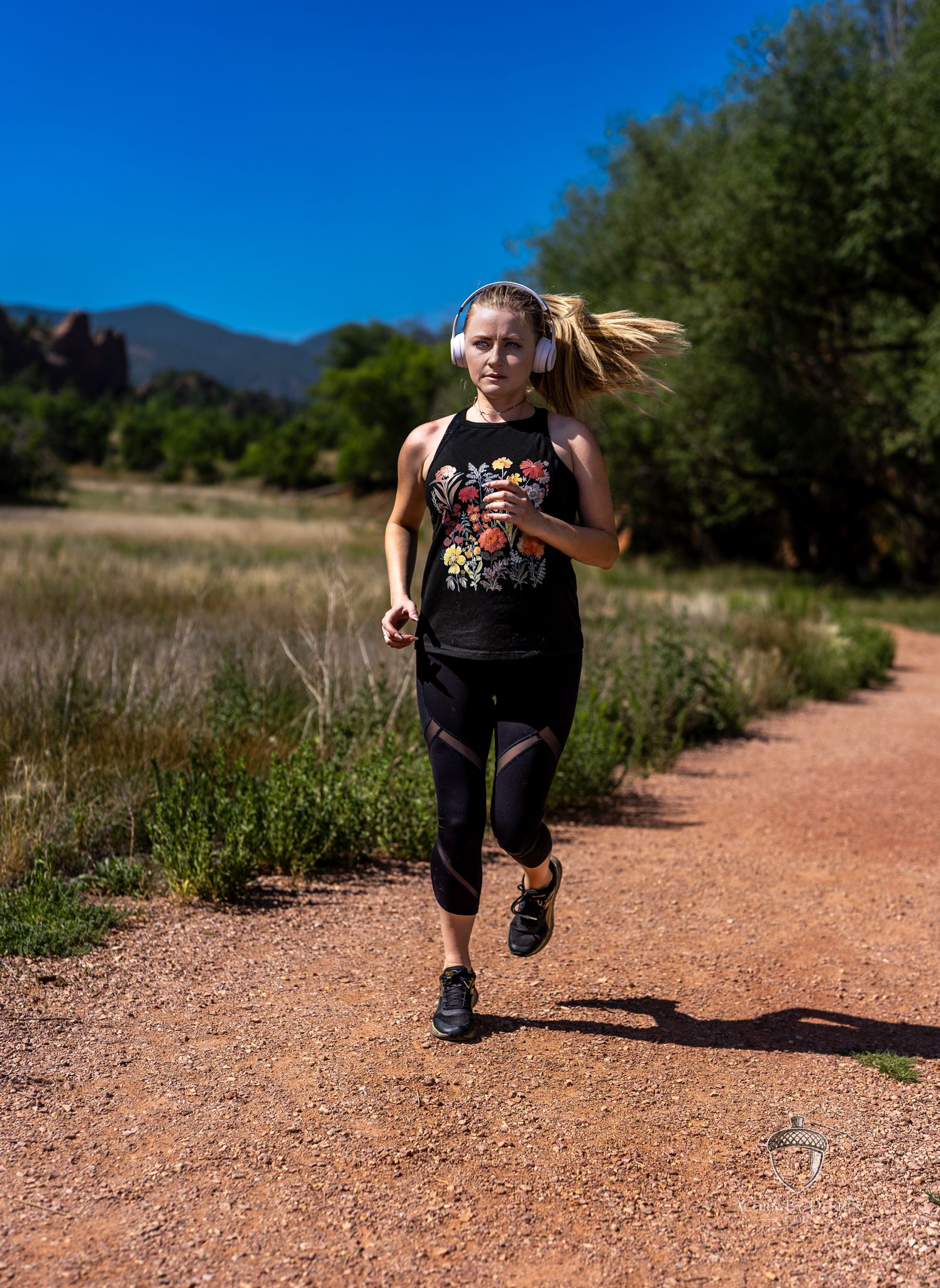 Woman running on a dirt trail wearing black workout clothes and white headphones with mountains in the background.