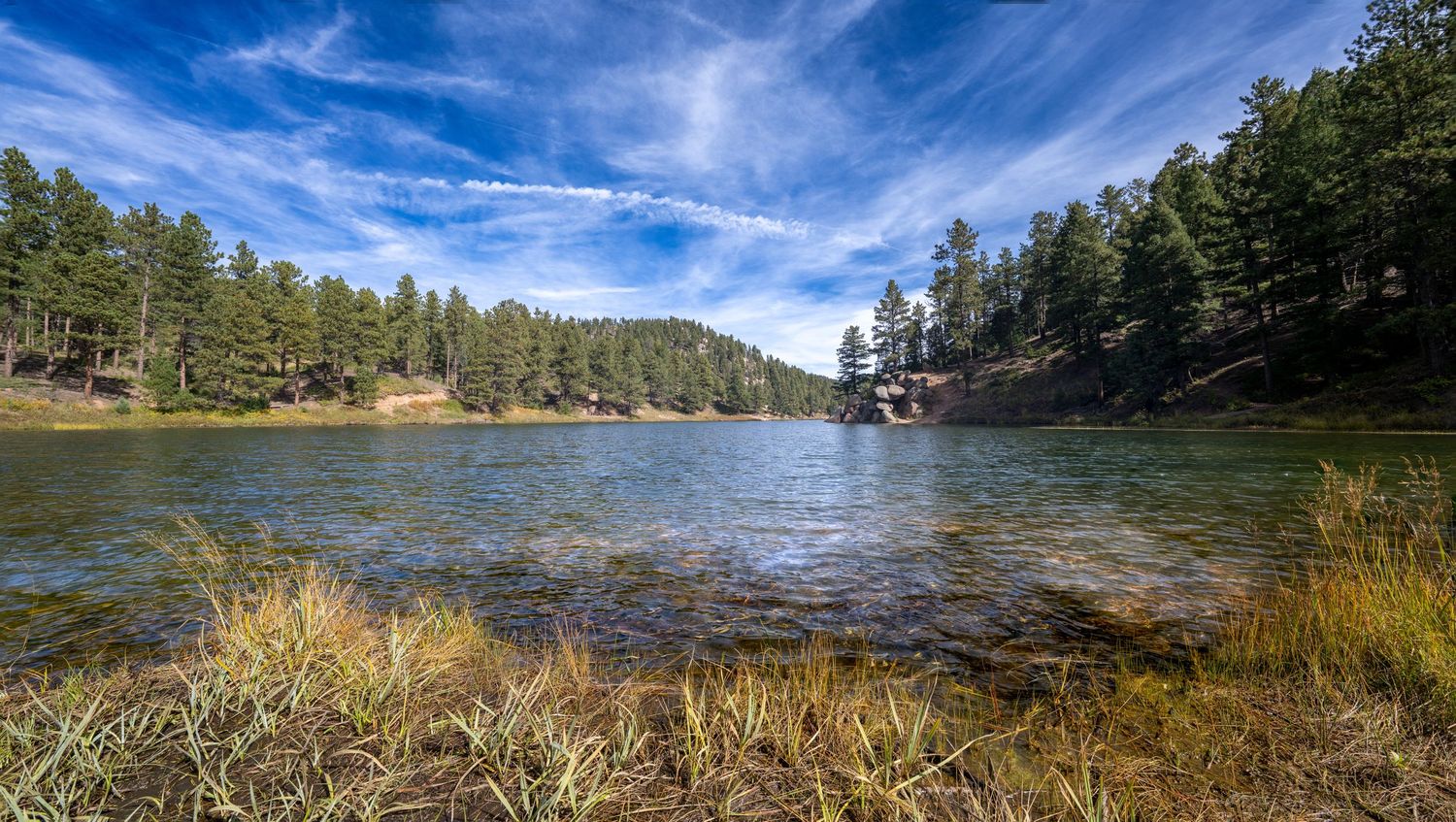 Scenic lake view surrounded by pine forest under bright blue summer sky with wispy clouds.