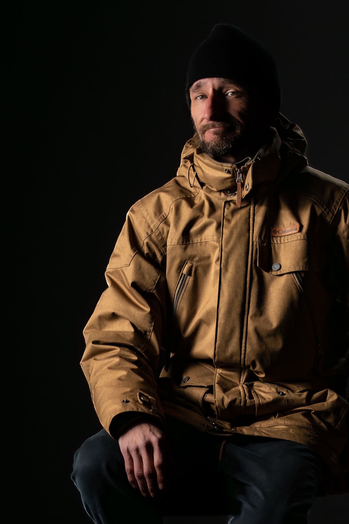 Man in yellow jacket and black beanie hat posing against dark background in dramatic studio lighting.