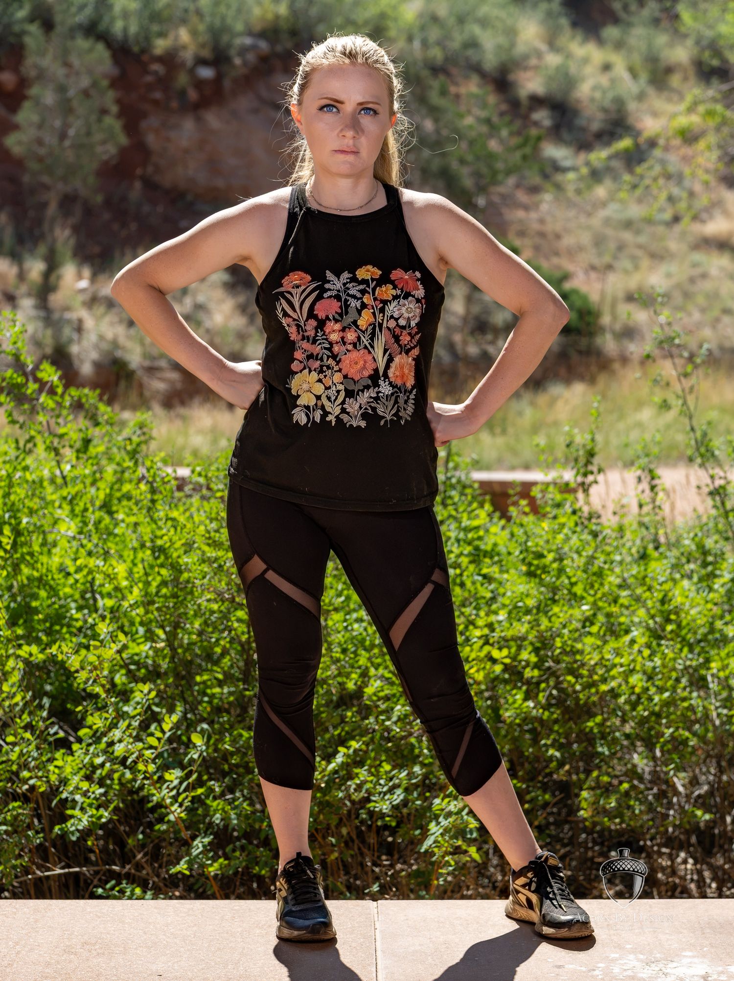 Woman in black floral tank top and mesh capri leggings poses outdoors with hands on hips, wearing athletic shoes.