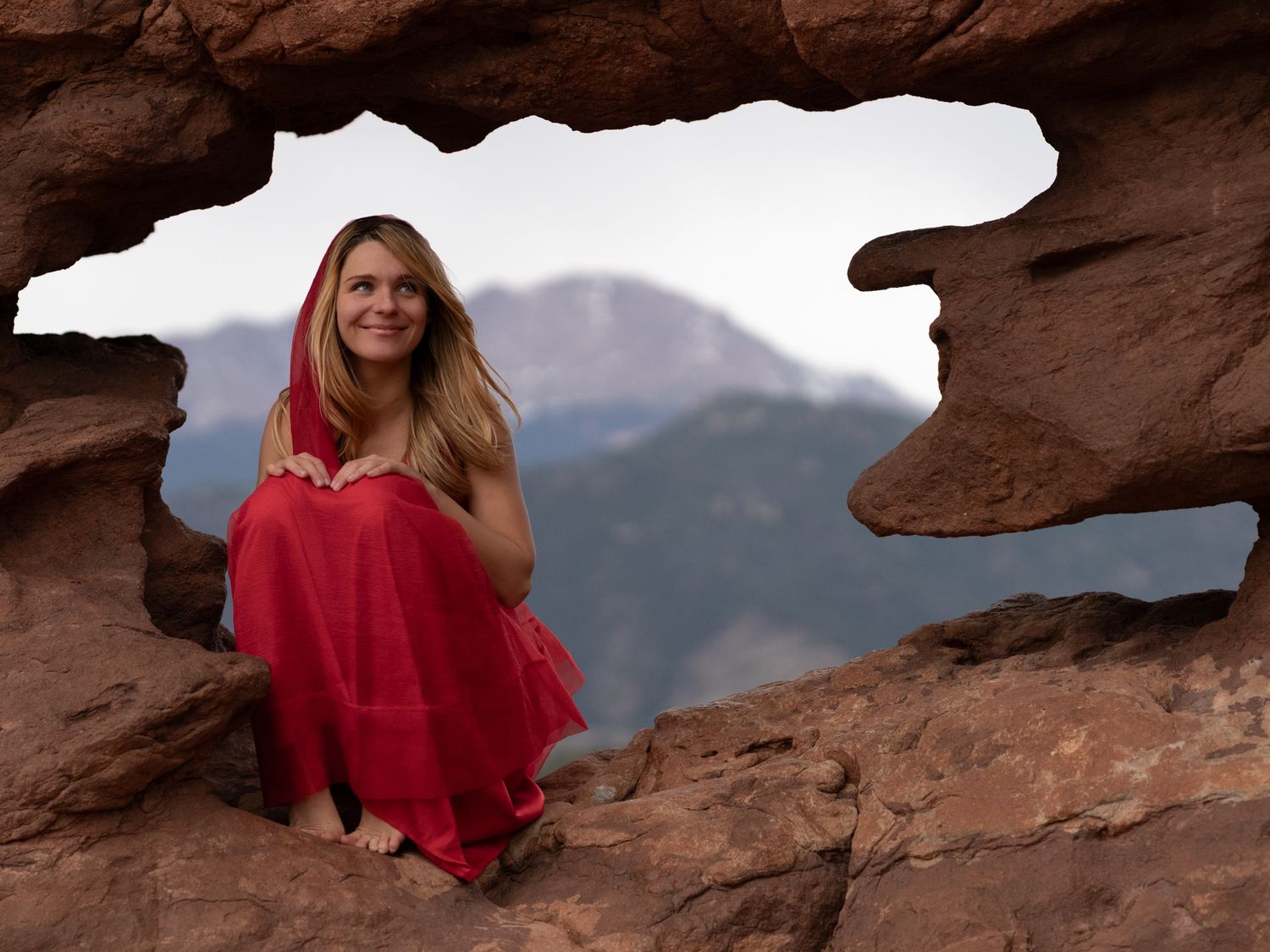 Woman in red dress sitting in red rock formation window with mountain backdrop.