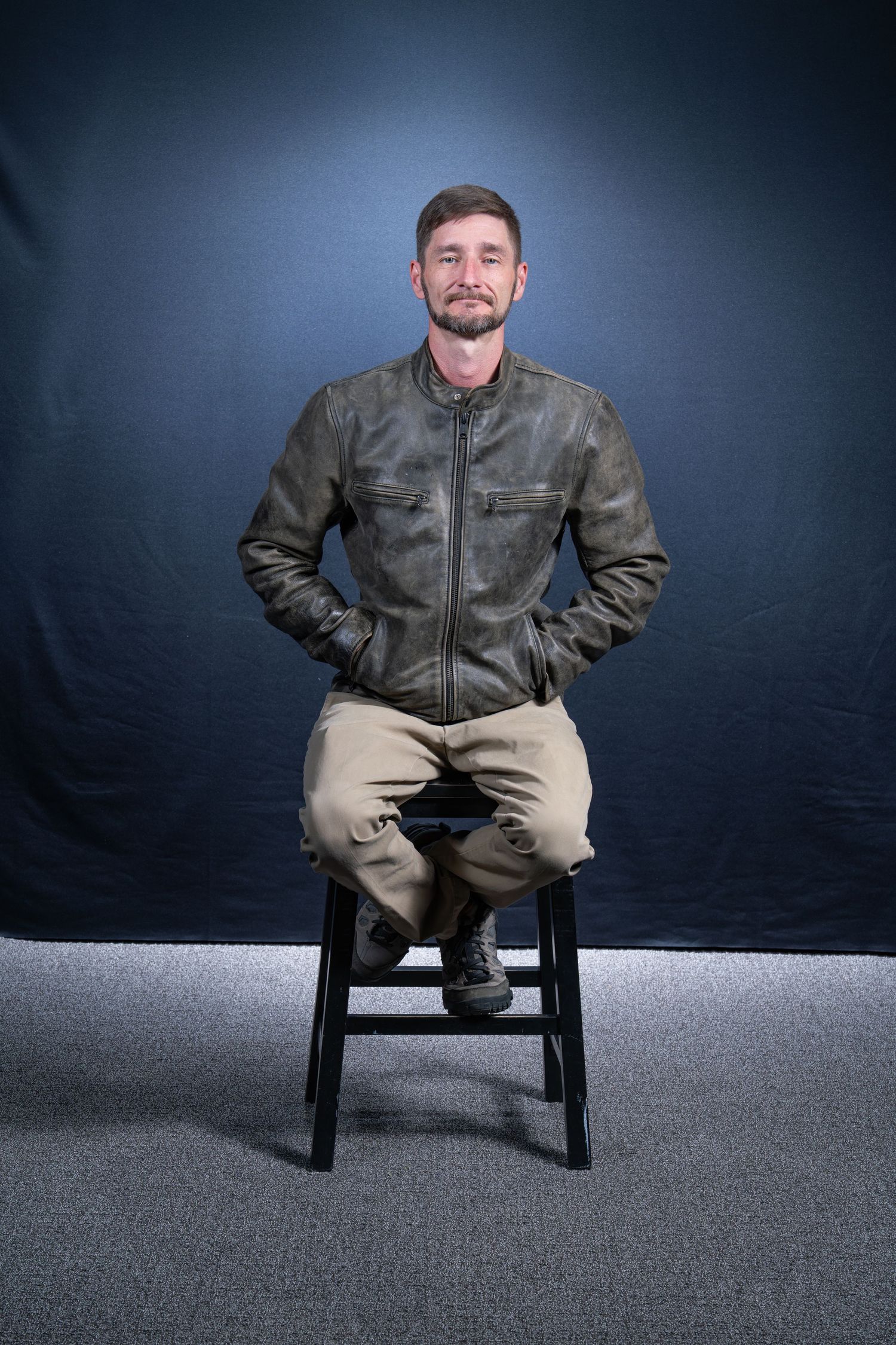 Person in leather jacket sitting on black chair against dark blue backdrop.