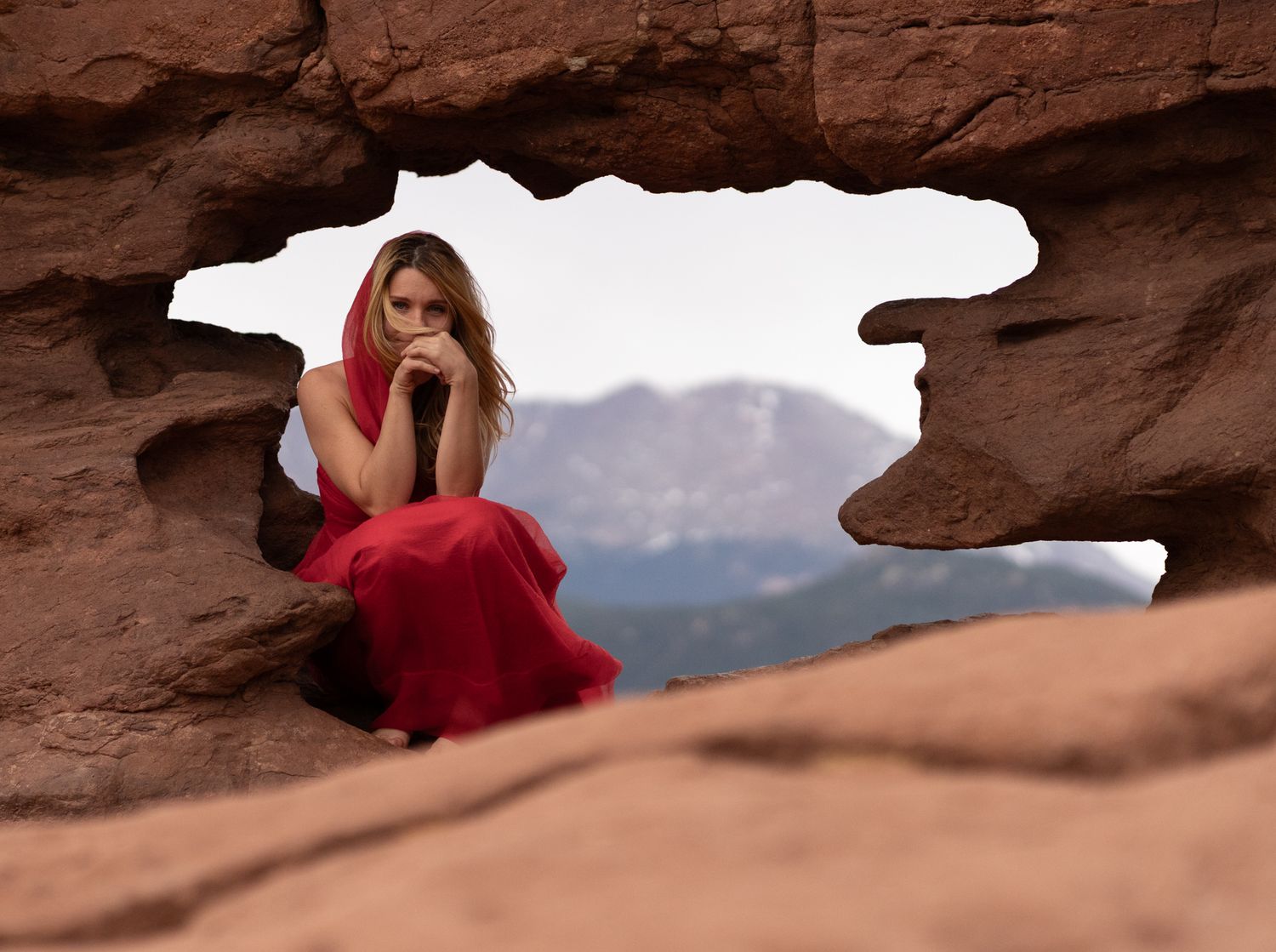 Woman in red dress sitting in a red rock arch opening with mountain views in Garden of the Gods.