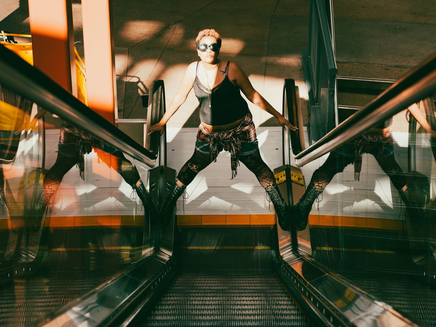 A punk-styled person poses dramatically on an escalator wearing dark clothing, boots, and sunglasses in a modern building.