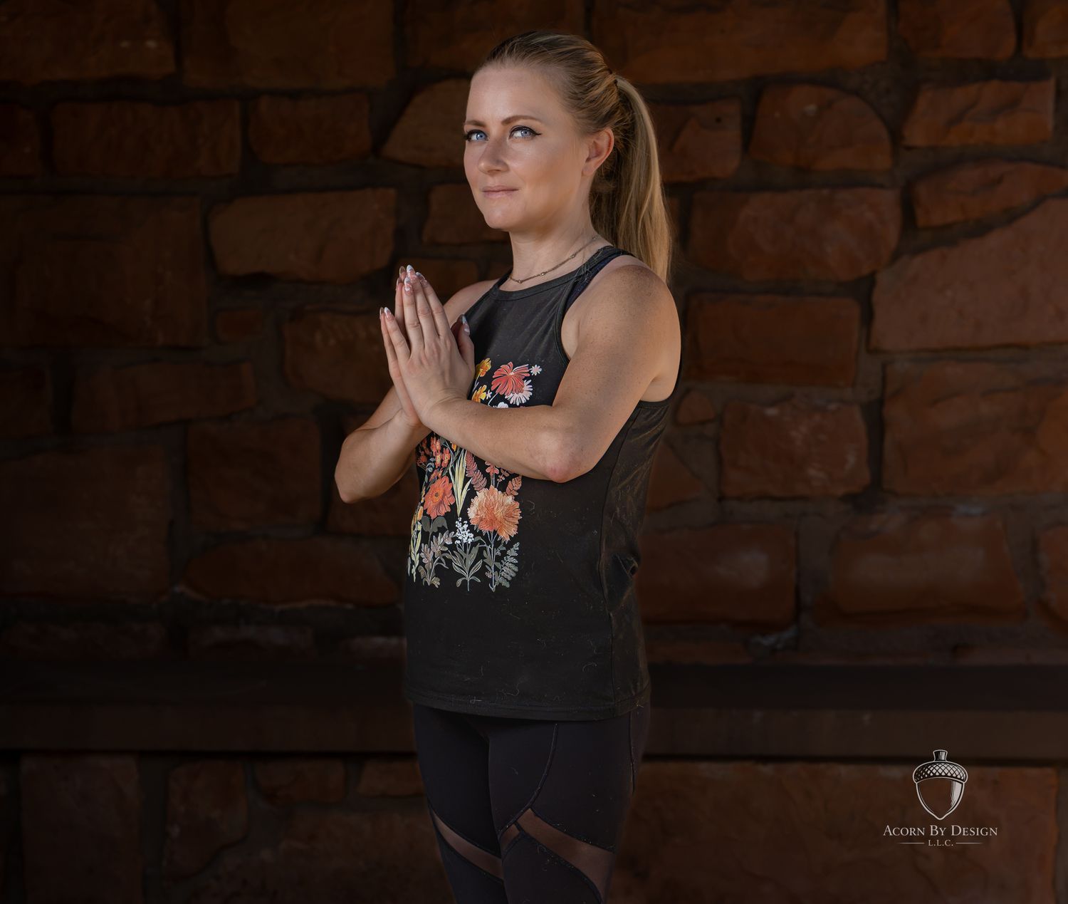 Woman in black floral tank top practicing yoga prayer pose against rustic stone wall background.