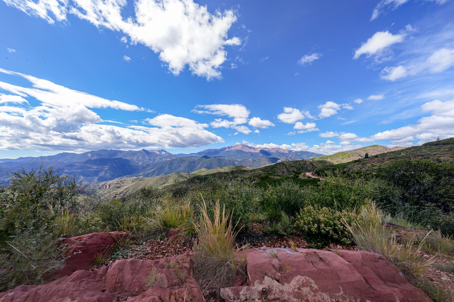Panoramic mountain landscape with dramatic clouds and red rock formations in the distance.