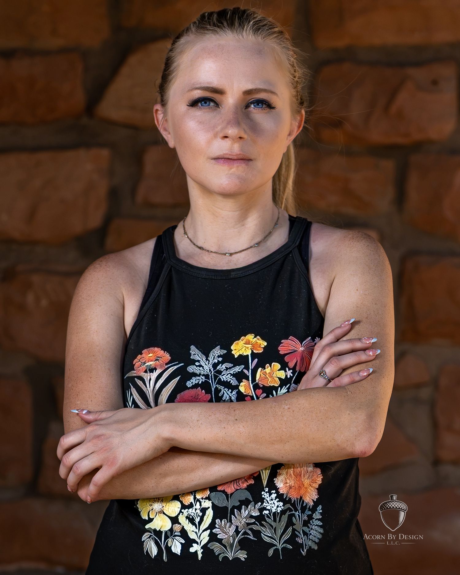 Young woman with blonde hair, arms crossed, wearing a black floral tank top, posing against a stone wall backdrop.