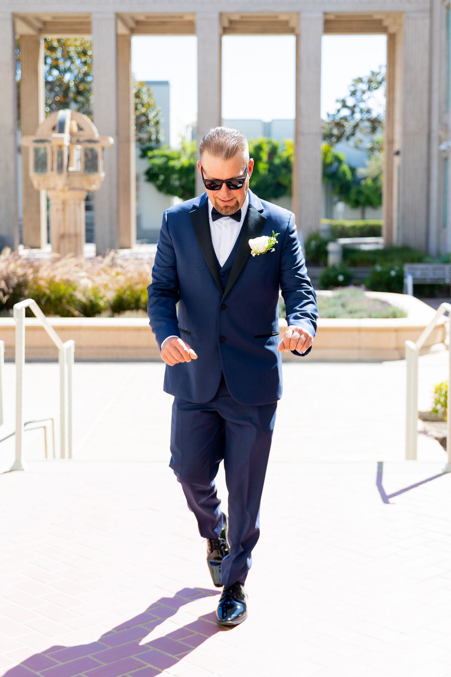 Groom in navy blue tuxedo with bow tie and boutonniere walking confidently outdoors on a sunny wedding day.