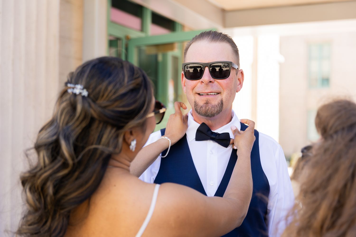 Bride adjusting groom's bow tie outdoors, groom wearing sunglasses and navy vest with suspenders on wedding day.