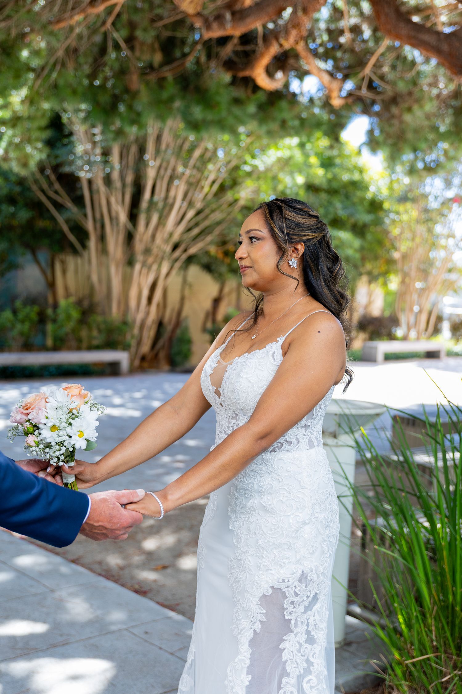 Bride in lace wedding gown holds bouquet of white flowers while holding hands with groom outdoors.