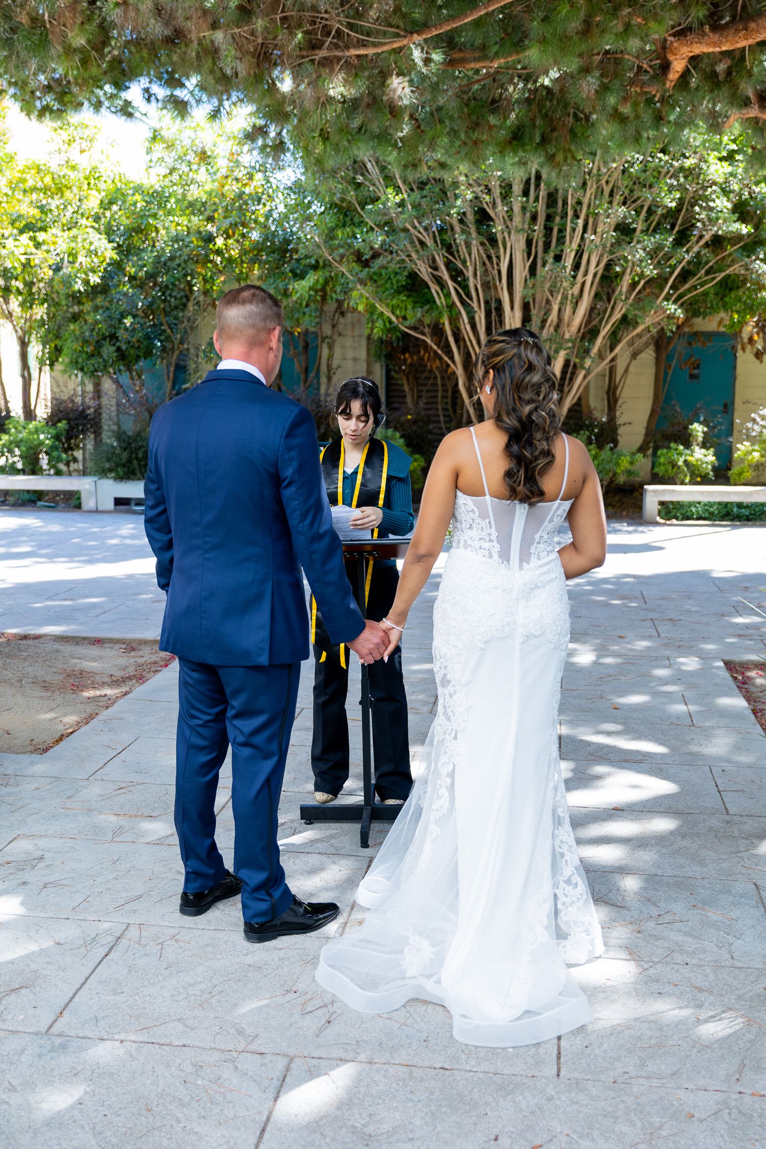 Bride and groom hold hands during outdoor wedding ceremony with officiant in green robe amid lush garden setting.