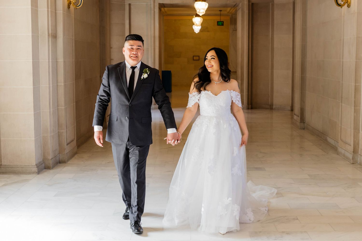 A bride and groom walk hand in hand through an elegant hallway with marble columns and warm lighting.