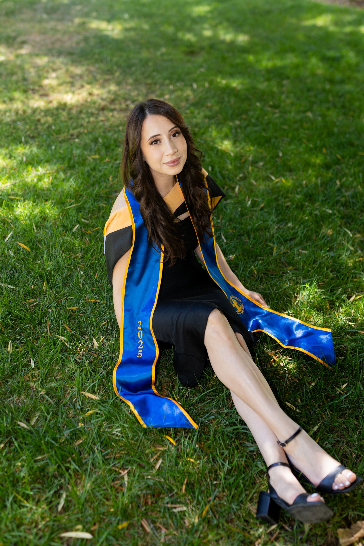 Young woman in black dress and blue gold graduation sash sits on green grass for outdoor graduation portrait.