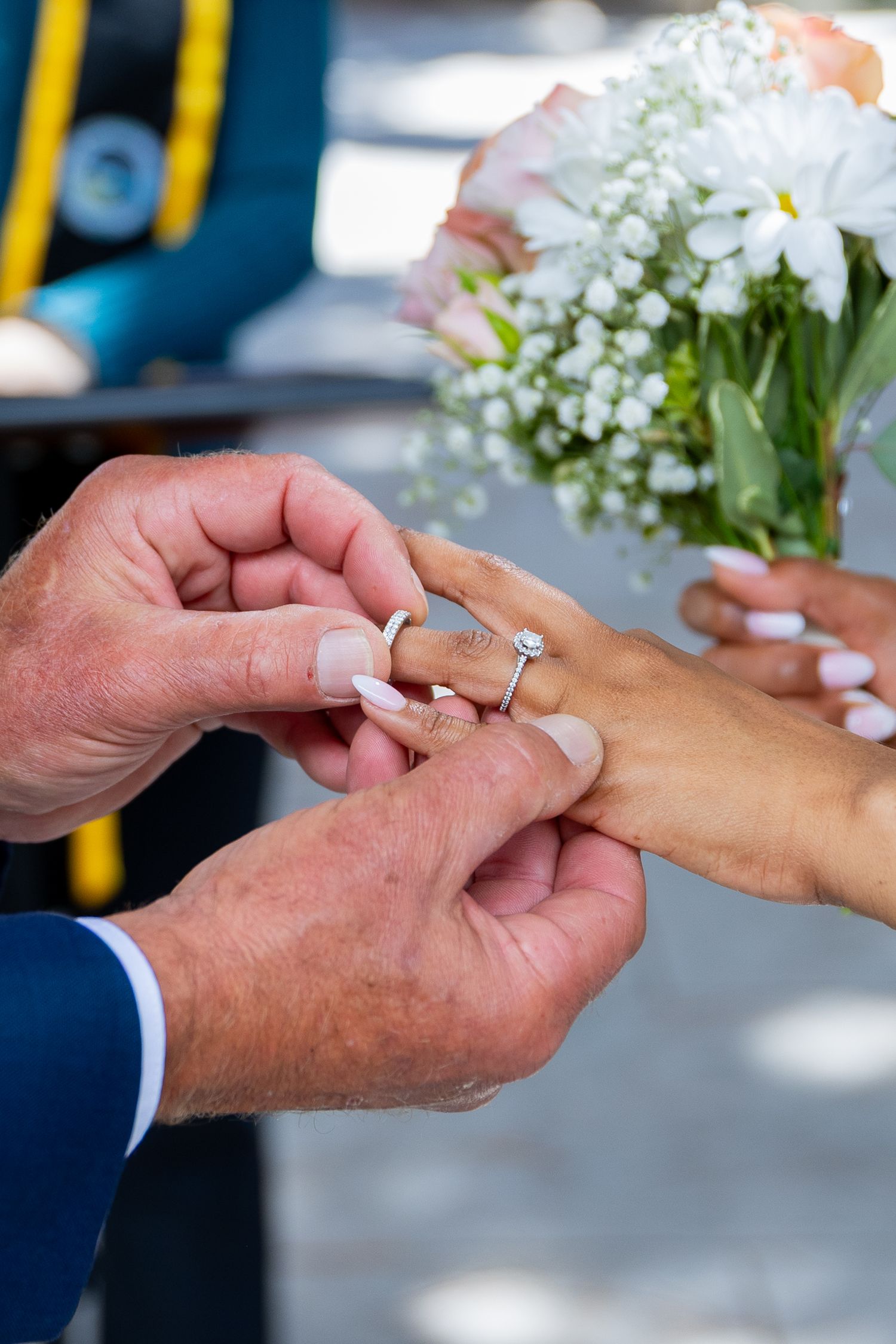 Groom placing a diamond wedding ring on bride's finger during ceremony, with bridal bouquet of white daisies in background.