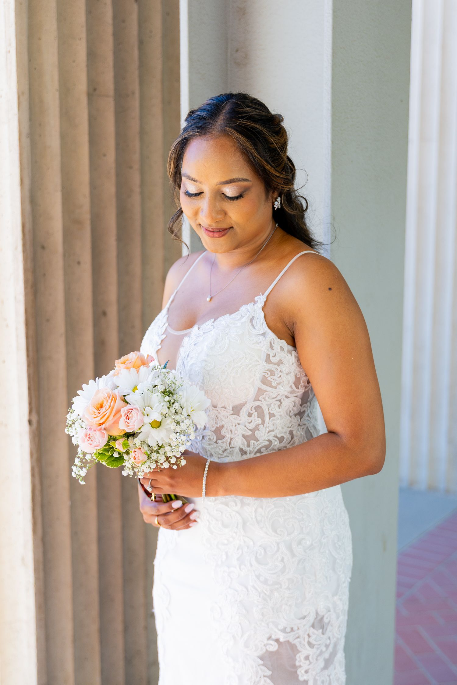 Bride in white lace gown holding peach and white bouquet, standing by stone columns, looking down gracefully.