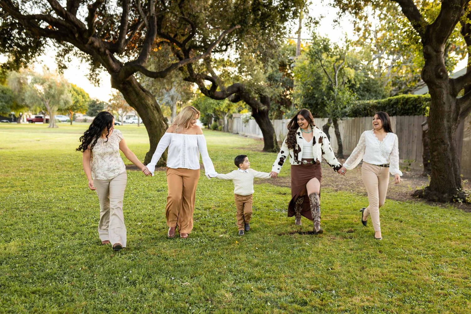 Five women and a child hold hands walking through a sunny park with large oak trees on green grass.