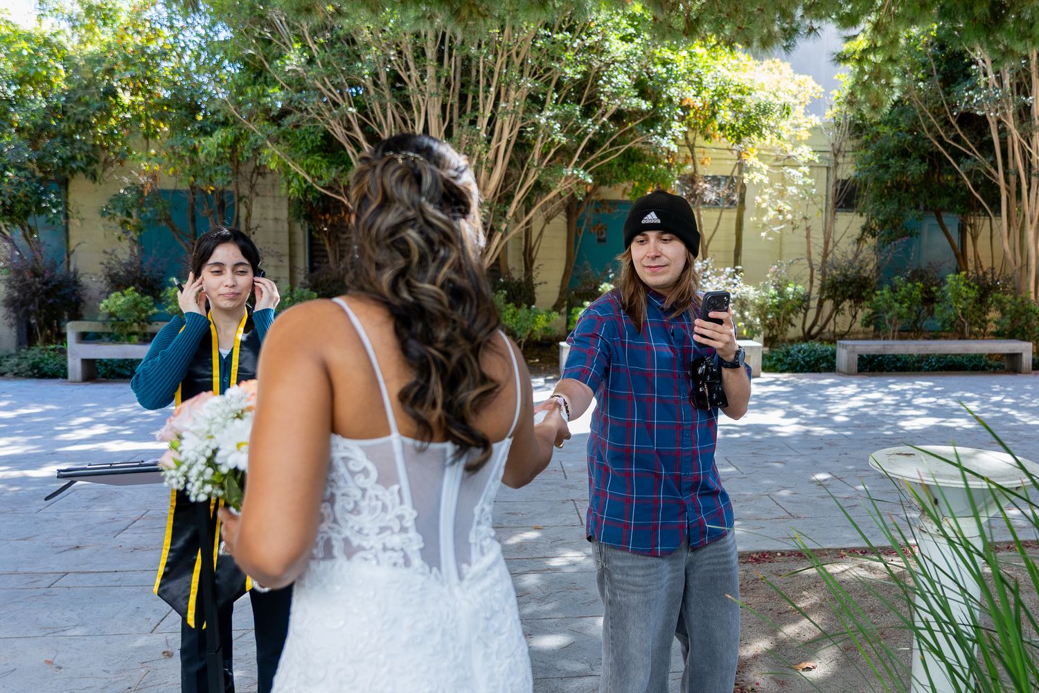 Bride in white lace dress holds flowers as two guests photograph her outdoors at a garden wedding venue.