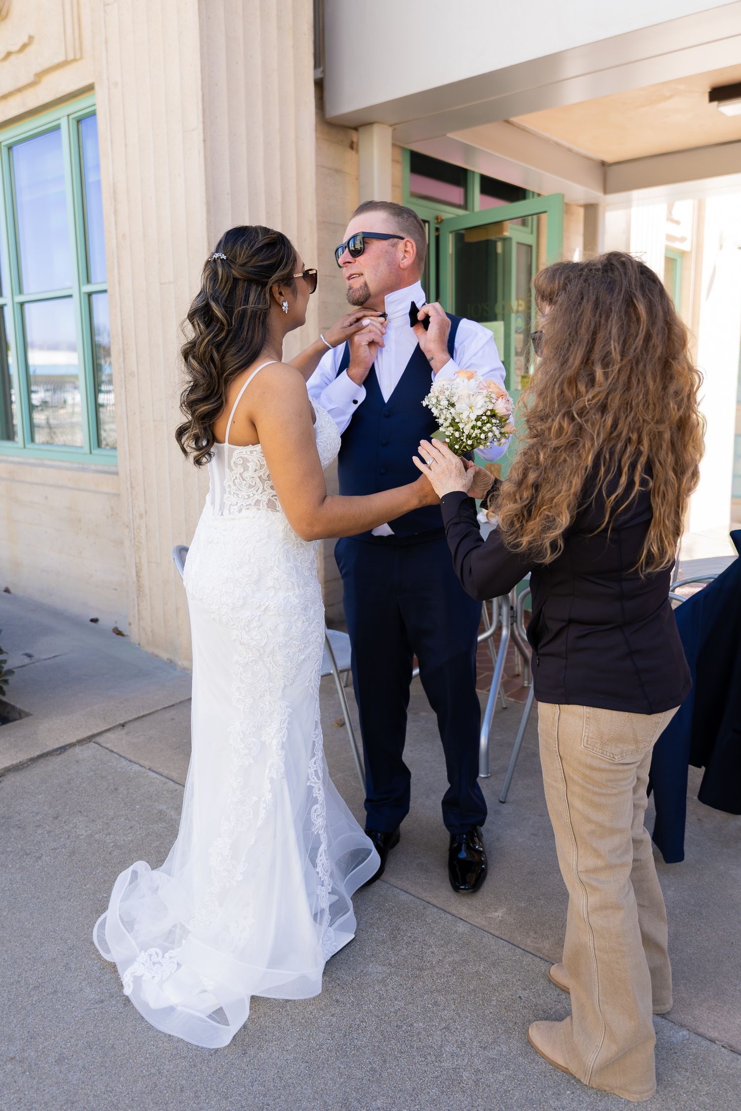 Bride in white lace gown adjusting groom's tie outdoors before wedding ceremony.