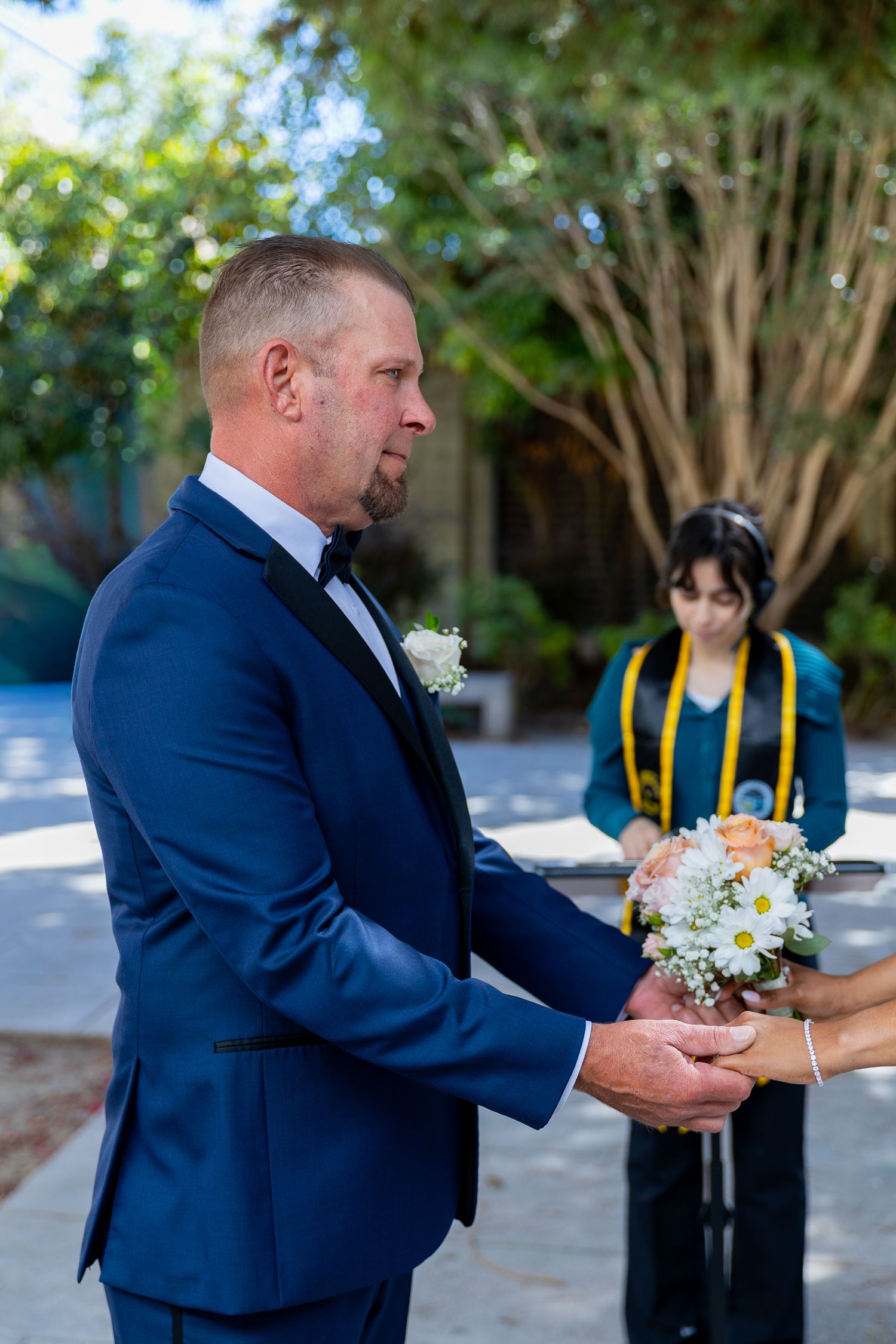 A groom in a navy blue suit holds a bridal bouquet of white and pink flowers during an outdoor wedding ceremony.
