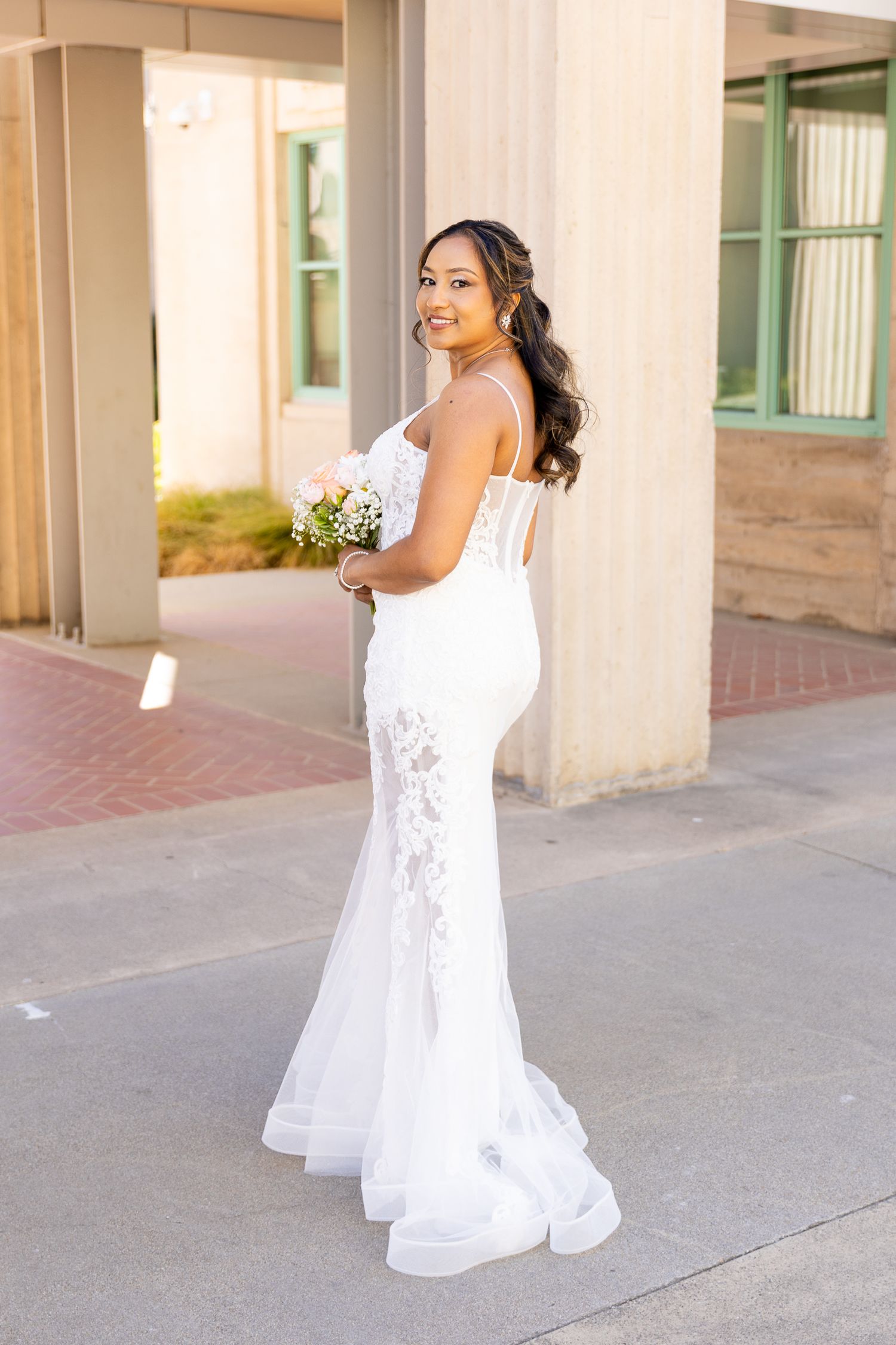 Smiling bride in white lace mermaid gown holding pink bouquet, posing outdoors near building columns.