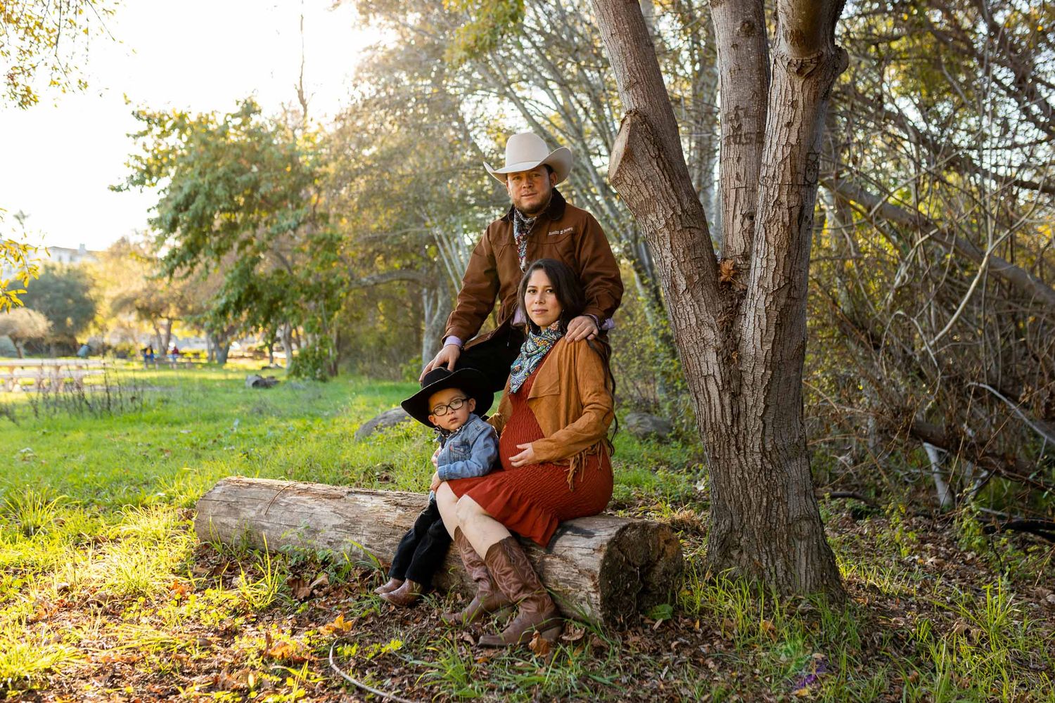Family of three in western attire poses by a tree on a log in a sunlit green field.