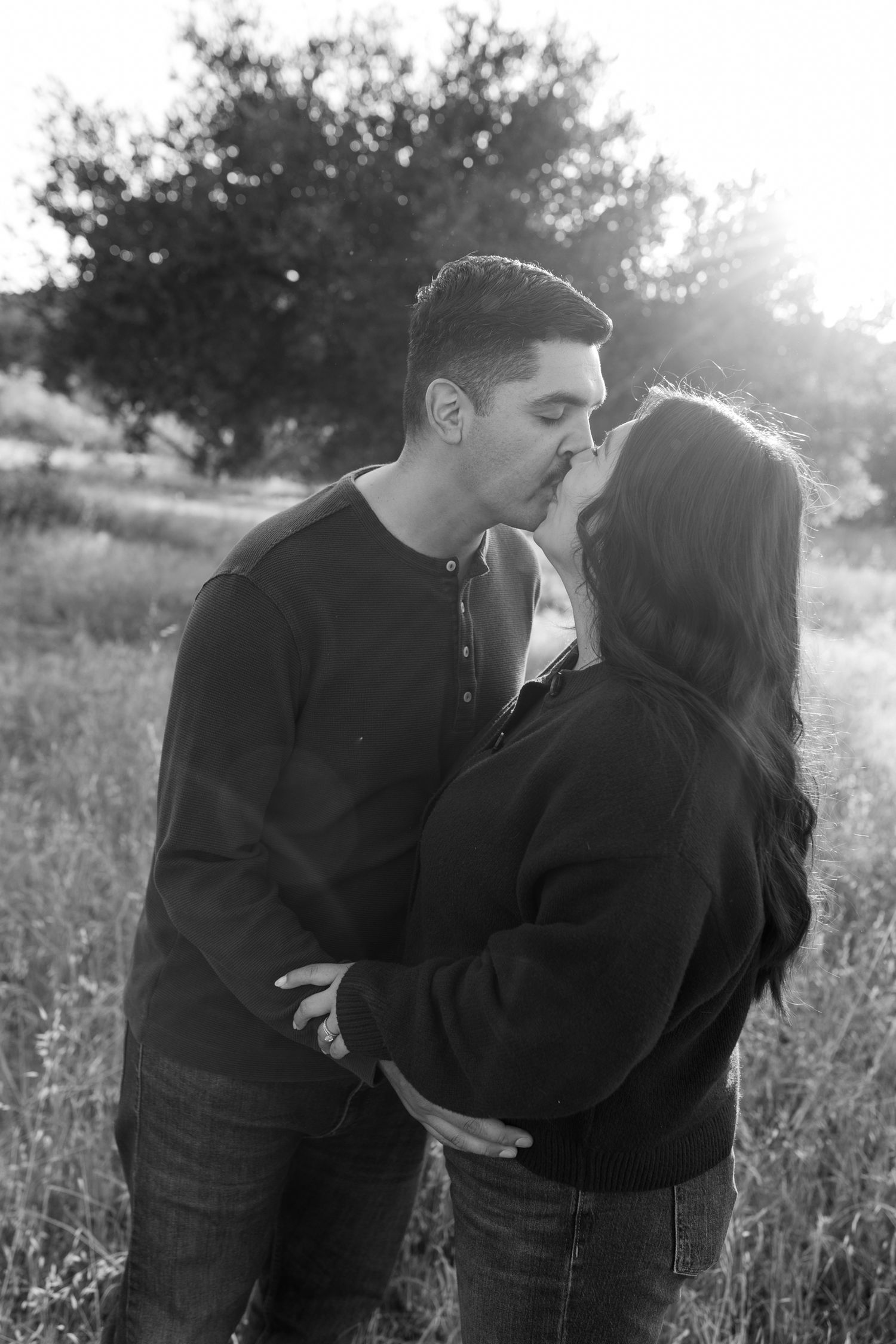 Black and white photo of a couple kissing in a sunlit field with trees in the background.