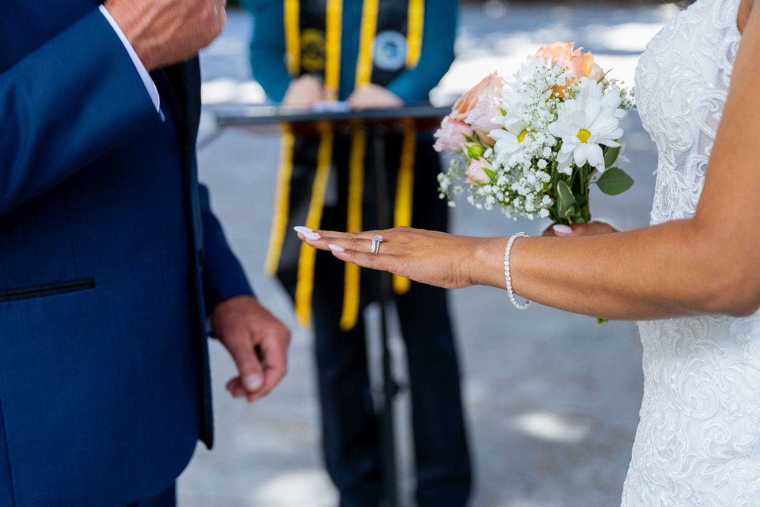 Bride holding white daisy bouquet while groom places wedding ring on her finger during outdoor ceremony.